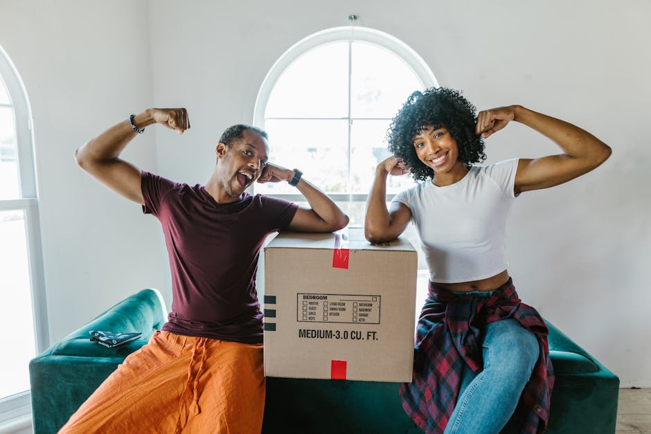 A man and woman inside a bright room with white walls and a large arched window in the background, posing with flexed arms near a medium-sized cardboard moving box labeled with room details and volume