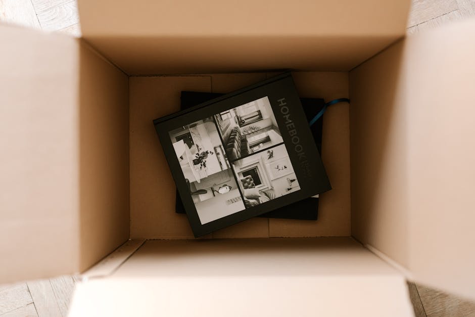 A man and woman inside a bright room with white walls and a large arched window in the background, posing with flexed arms near a medium-sized cardboard moving box labeled with room details and volume