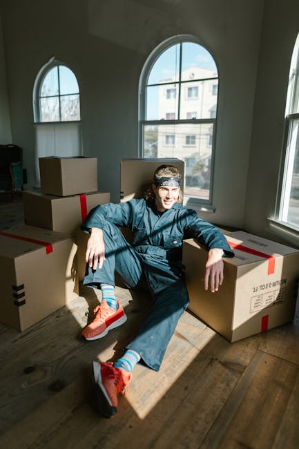 A young man with long hair, wearing a navy blue jumpsuit, bright orange running shoes, and striped socks, sits on a wooden floor inside a room near three large cardboard boxes sealed with red tape. Th