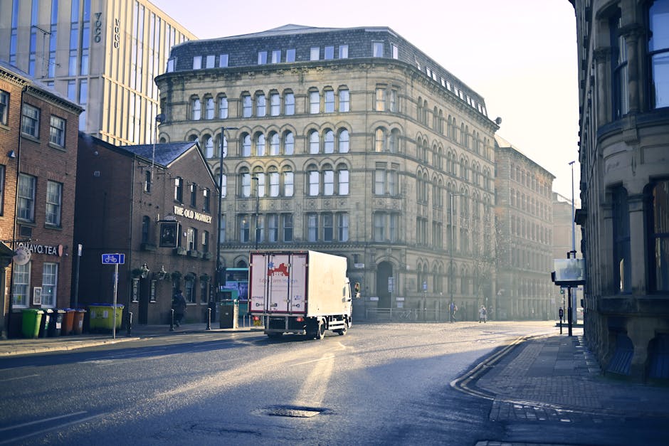 A street scene in Roehampton during daylight shows a white moving truck with a box on its back, positioned in front of an ornate multi-storey building with arched windows. The truck is parked on a pav
